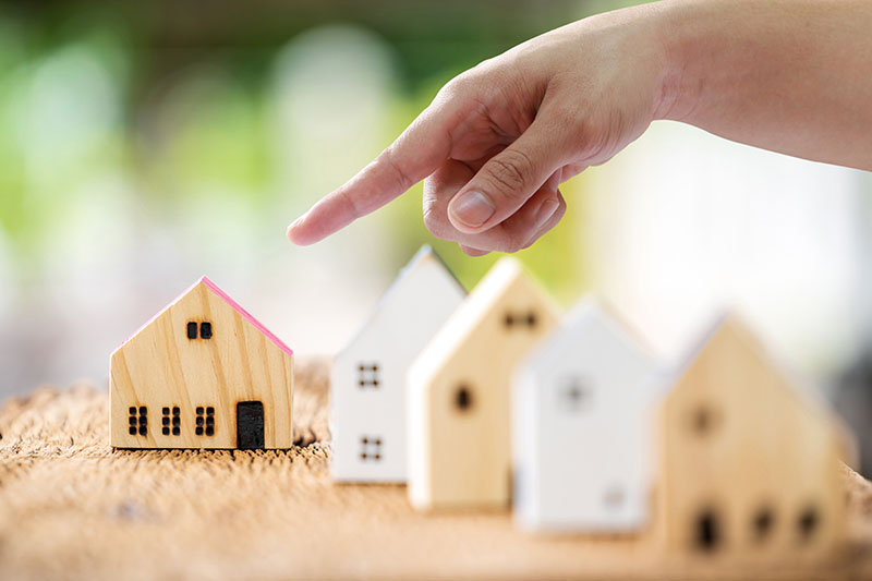 A hand points toward a small wooden house model with a pink roof among other similar house models placed on a textured surface, suggesting the concept of choosing a home or property selection.
